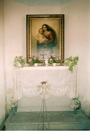 Na zdi je zavěšen velký obraz Panny Marie           chovající dítě, pod ním na dřevěném schodu stojí bíle prostřený stůl           s umělými květinami, křížem, dvěma svícni a opět malou bílou soškou           Panny Mariie. Před stolem je bílá svíce ve svícnu na třech vysokých           nohách
