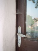 Detail na zakulacenou kliku s orezlými šrouby, hnědé dveře a futra,         která jsou zakryta pavučinami, ve skle se leskne nedostavěný dům