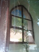 Pohled srkze sklo dvěří na okno zakončené lomenným obloukem         rozdělené na šest dílů. Levý prostřední je otevírací, na jehož kličce         visí uvýzlý ztrouchnivělé lano od zvonu. Tabulka levého horního dílu         okna je popraskaná, vrchní tabulky ukazují modrou plachtu, prostřední         zelené listí a spodní bílý dům se červenými taškami. Ve skle dvěří přes         které blo foceno se odráží vesnická chalupa bílo-okrové omítky se starou         červenou střechou, před domem je hnědý plot, z části řidký, z části         hustý a na předzahrádce roste vysoký smrk.